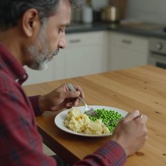 A man in his late sixties eating a plate of mashed potatoes and peas at a table