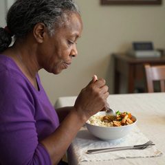 A woman in her late sixties eating a rice bowl with chicken and vegetables at a table