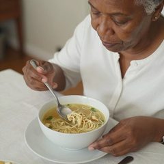A woman in her seventies eating a bowl of chicken noodle soup at a table