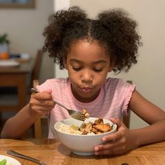 A girl about seven years old eating a rice bowl with chicken and vegetables at a table