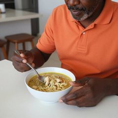A man in his seventies eating with one hand eating a bowl of chicken noodle soup at a table