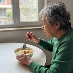 A woman in her late sixties eating a bowl of chicken noodle soup at a table