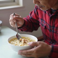 A man in his seventies eating a bowl of chicken noodle soup at a table