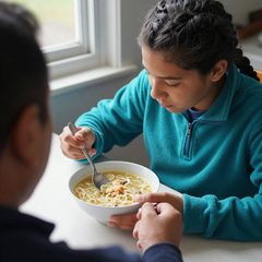 A parent and a teenager eating a bowl of chicken noodle soup at a table