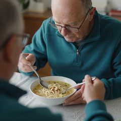 A man in his seventies eating a bowl of chicken noodle soup at a table