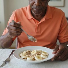 A man in his seventies eating a bowl of oatmeal with sliced banana at a table