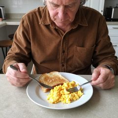 A man in his late sixties eating a plate of scrambled eggs and toast at a table